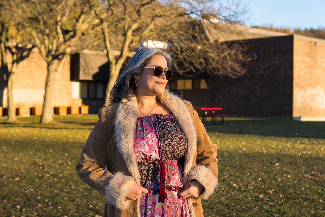 Cindy looking to the side in her suede fur-trimmed coat and multi patterned dress with lots of pinks and burgundies. She’s wearing aviators and has her hair down.