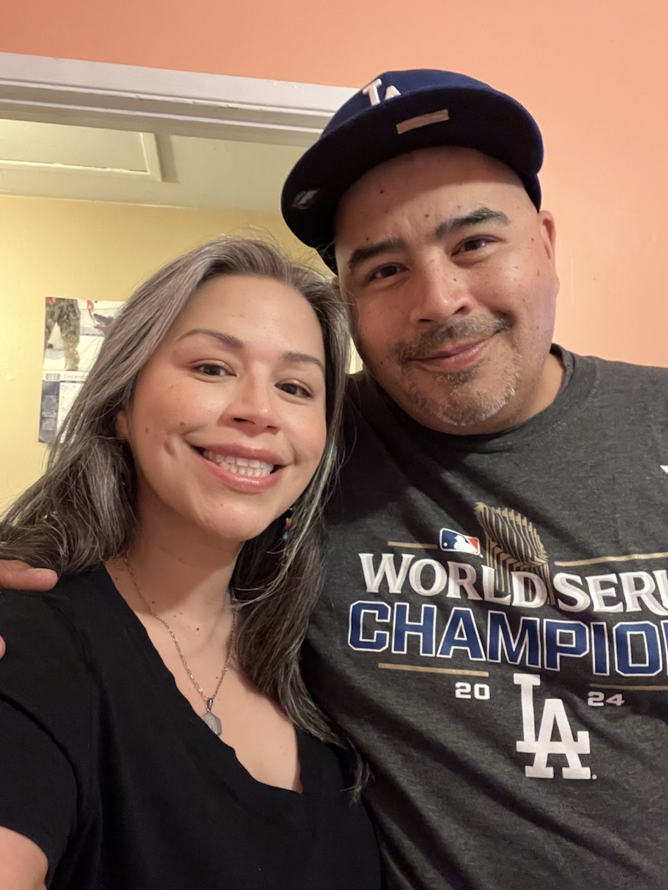 Cindy and her brother Danny side by side. They’re smiling at the camera. She’s in a black shirt and he’s in a grey LA Dodgers shirt and blue LA cap. 